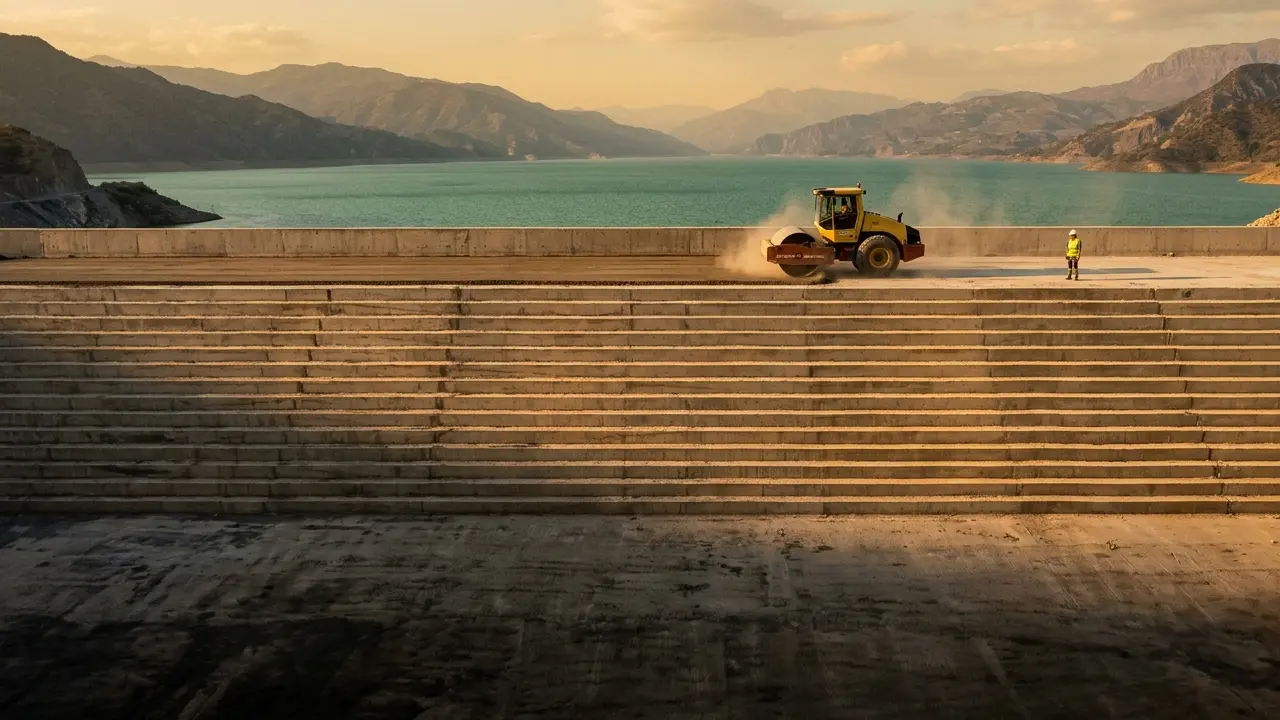 Golden-hour photo of a roller-compacted concrete (RCC) dam under construction: a yellow vibratory roller compacts a fresh RCC lift on the crest above the stepped downstream face, with a worker for scale and a teal-green reservoir behind.