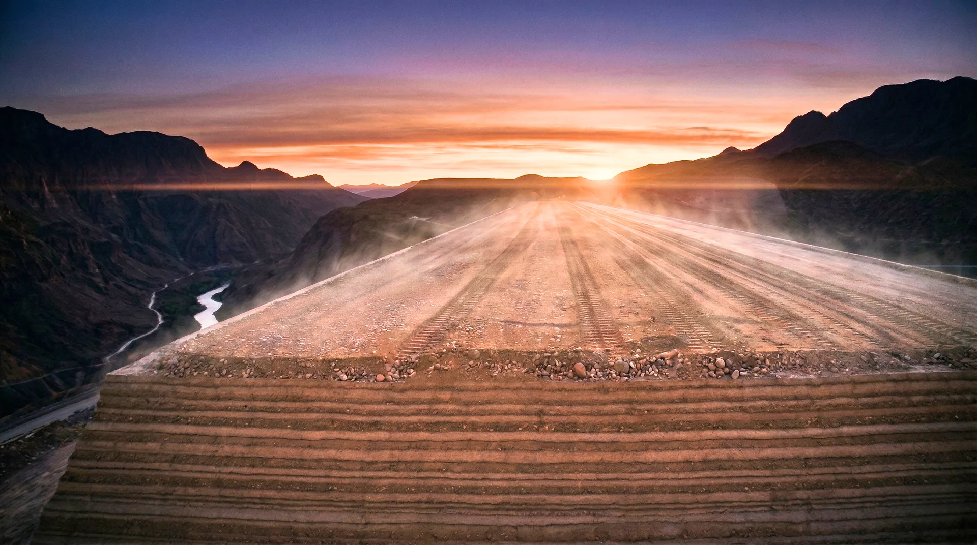 Aerial view of an RCC dam under construction at sunset, showing freshly compacted roller compacted concrete lifts with visible horizontal layer striations, heat shimmer rising from the surface, and a river gorge with mountain silhouettes in the background